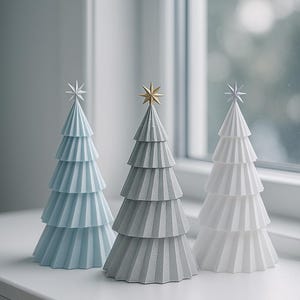 Op de afbeelding: Drie decoratieve papieren kerstbomen in lichtblauw, grijs en wit, elk met een ster als top. De bomen zijn gelaagd en staan op een witte vensterbank, wat een feestelijke sfeer creëert.