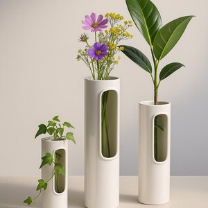 Op de afbeelding: Drie witte keramische vazen van verschillende hoogtes, elk met een rechthoekige uitsparing. De vazen bevatten groen en kleurrijke bloemen, waaronder paarse cosmos en gele bloemen. De opstelling staat tegen een neutrale achtergrond.