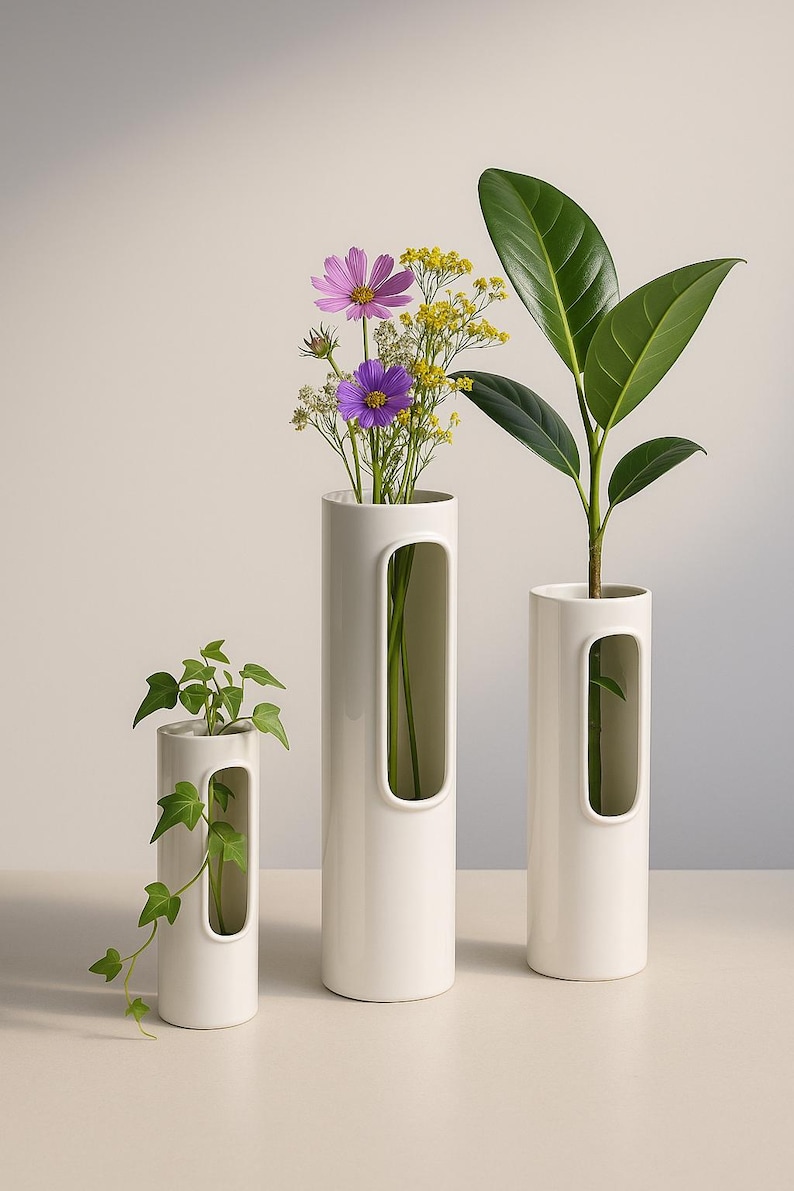 Op de afbeelding: Drie witte keramische vazen van verschillende hoogtes, elk met een rechthoekige uitsparing. De vazen bevatten groen en kleurrijke bloemen, waaronder paarse cosmos en gele bloemen. De opstelling staat tegen een neutrale achtergrond.