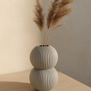 May include: A grey, ribbed ceramic vase with a stacked, spherical design, holding several dried, feathery brown stems. The vase sits on a light-coloured wooden surface, with a neutral-toned wall in the background.