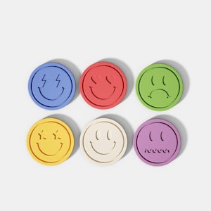 May include: Six colorful, round coasters with different smiley face designs. The coasters are blue with lightning bolts, red with hearts, green with a frown, yellow with stars, off-white with a smile, and purple with a grimace.