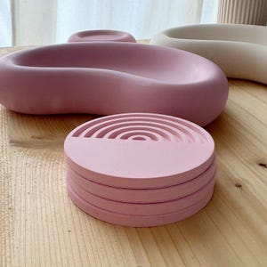 Puede incluir: Una pila de posavasos redondos rosas con un diseño de arcoíris, junto con una bandeja rosa grande y curvada y otras piezas decorativas. Los artículos están hechos de un material sólido y se encuentran sobre una superficie de madera.
