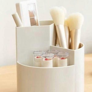 May include: A white, multi-compartment makeup organizer filled with various beauty products. The organizer holds makeup brushes, lipsticks, and an eyeshadow palette. The organizer is made of a smooth, solid material and is designed to keep cosmetics organized.
