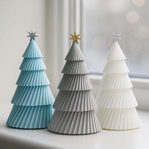 Könnte beinhalten: Drei dekorative Weihnachtsbäume in Blau, Grau und Weiß. Jeder Baum hat ein geschichtetes, konisches Design mit einem Stern oben. Die Bäume werden auf einer weißen Oberfläche in der Nähe eines Fensters ausgestellt.