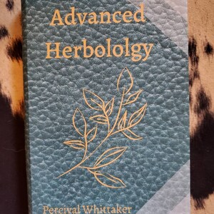 May include: A teal green book with a gold embossed title "Advanced Herbology" and a gold illustration of a leafy branch. The author's name, Percival Whittaker, is printed in gold at the bottom of the cover.