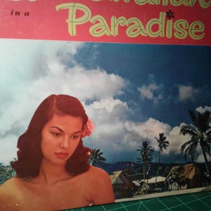 May include: Vintage vinyl record album cover titled "Hawaiian Paradise." The cover features a woman with dark hair and a pink flower, set against a tropical landscape with palm trees and a blue sky.