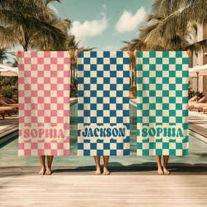Op de afbeelding: Drie strandlakens met een geblokt patroon in roze, blauw en teal. Elke handdoek heeft een naam onderaan gedrukt: "SOPHIA" in roze, "JACKSON" in blauw en "SOPHIA" in teal. De handdoeken worden voor een zwembad gehouden.