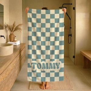Op de afbeelding: Een grote, geblokte badhanddoek in cr&egrave;me en lichtblauw, met de naam "TOMMY" in een retro lettertype. De handdoek wordt getoond in een badkamer, waarbij het ontwerp en de kleurstelling worden benadrukt.