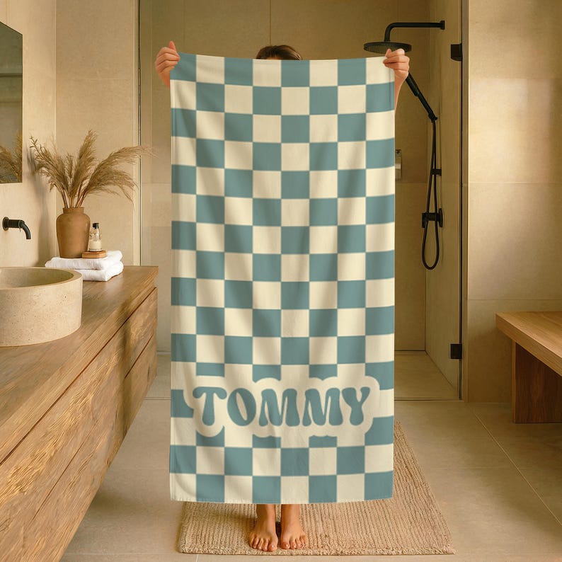 Op de afbeelding: Een grote, geblokte badhanddoek in cr&egrave;me en lichtblauw, met de naam "TOMMY" in een retro lettertype. De handdoek wordt getoond in een badkamer, waarbij het ontwerp en de kleurstelling worden benadrukt.