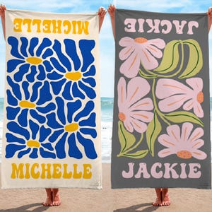 K&ouml;nnte beinhalten: Zwei Strandt&uuml;cher mit floralen Designs. Eines ist cremefarben mit blauen Blumen und dem Namen "MICHELLE" in Gelb. Das andere ist grau mit rosa Blumen und dem Namen "JACKIE" in Rosa. Beide werden am Strand gehalten.