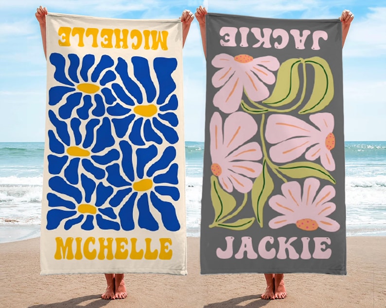 K&ouml;nnte beinhalten: Zwei Strandt&uuml;cher mit floralen Designs. Eines ist cremefarben mit blauen Blumen und dem Namen "MICHELLE" in Gelb. Das andere ist grau mit rosa Blumen und dem Namen "JACKIE" in Rosa. Beide werden am Strand gehalten.