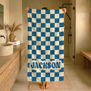 Op de afbeelding: Een blauw en cr&egrave;me geruite badhanddoek met de naam "JACKSON" in blauwe letters. De handdoek wordt in een badkameromgeving omhoog gehouden, waarbij het ontwerp en de kleurstelling worden getoond. De handdoek is rechthoekig en lijkt van zacht materiaal te zijn.