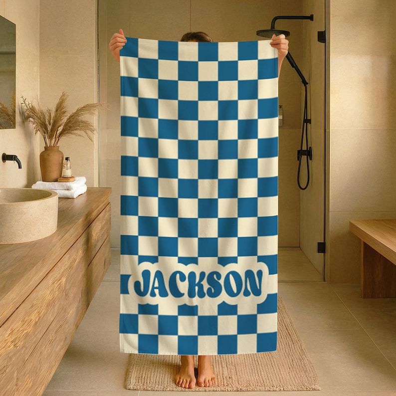 Op de afbeelding: Een blauw en cr&egrave;me geruite badhanddoek met de naam "JACKSON" in blauwe letters. De handdoek wordt in een badkameromgeving omhoog gehouden, waarbij het ontwerp en de kleurstelling worden getoond. De handdoek is rechthoekig en lijkt van zacht materiaal te zijn.