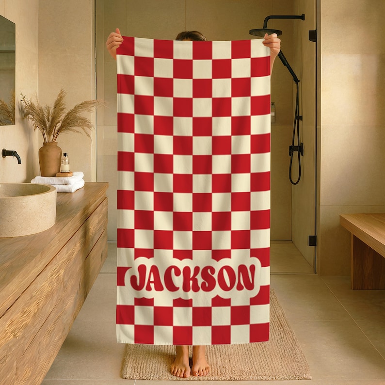 Op de afbeelding: Een rood en cr&egrave;me geblokte badhanddoek met de naam "JACKSON" in rode, gestileerde tekst. De handdoek wordt in een badkameromgeving omhoog gehouden. Het patroon van de handdoek is een herhalend raster van rode vierkanten tegen een cr&egrave;mekleurige achtergrond.