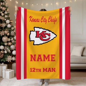 May include: A yellow, red, and white blanket featuring the Kansas City Chiefs logo and text. The blanket has a large yellow center with the team name and logo, flanked by red and white stripes. The text "NAME" and "12TH MAN" are also displayed.