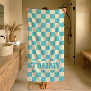 Op de afbeelding: Een grote, rechthoekige handdoek met een geblokt patroon in lichtblauw en cr&egrave;me. Het woord "TOMMY" staat in lichtblauw onderaan. De handdoek wordt in een badkamer gehouden.