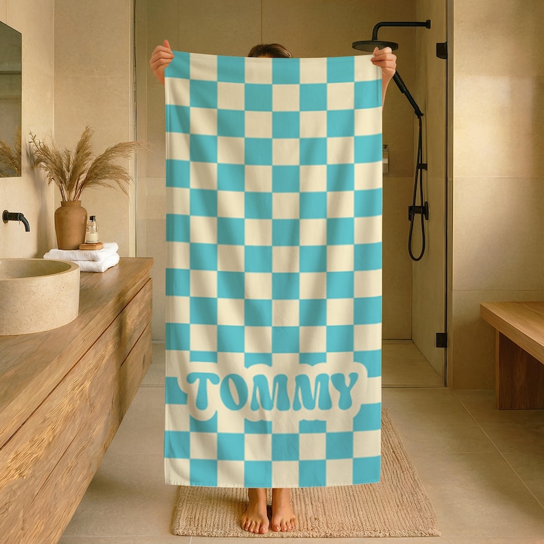 Op de afbeelding: Een grote, rechthoekige handdoek met een geblokt patroon in lichtblauw en cr&egrave;me. Het woord "TOMMY" staat in lichtblauw onderaan. De handdoek wordt in een badkamer gehouden.