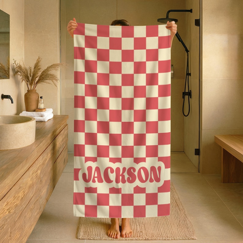 Op de afbeelding: Een rood en cr&egrave;mekleurig geblokte badhanddoek met de naam "JACKSON" in een retro lettertype. De handdoek wordt in een badkamer gehouden, waarbij het ontwerp en de kleurstelling worden getoond. Het patroon van de handdoek is een herhalend raster van rode en cr&egrave;mekleurige vierkanten.