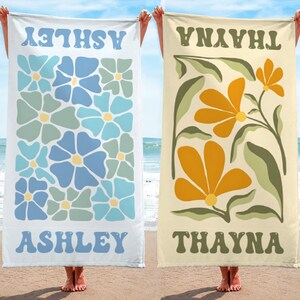 K&ouml;nnte beinhalten: Zwei Strandt&uuml;cher werden gezeigt, jedes mit einem Namen oben und unten. Eines ist wei&szlig; mit blau-gr&uuml;nen Blumenmustern und dem Namen "ASHLEY". Das andere ist beige mit orangefarbenen Blumenmustern und dem Namen "THAYNA".