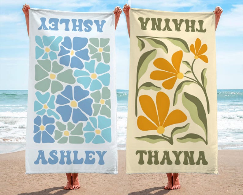 K&ouml;nnte beinhalten: Zwei Strandt&uuml;cher werden gezeigt, jedes mit einem Namen oben und unten. Eines ist wei&szlig; mit blau-gr&uuml;nen Blumenmustern und dem Namen "ASHLEY". Das andere ist beige mit orangefarbenen Blumenmustern und dem Namen "THAYNA".