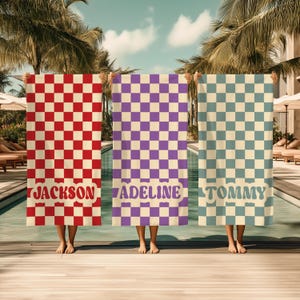 Op de afbeelding: Drie gepersonaliseerde strandlakens met dambordpatronen in rood, paars en blauw. Elk laken toont een naam in een retro lettertype: "JACKSON", "ADELINE" en "TOMMY". De lakens worden bij een zwembad omhoog gehouden.