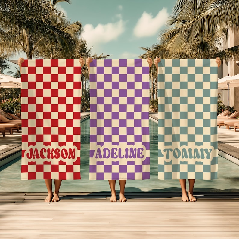 Op de afbeelding: Drie gepersonaliseerde strandlakens met dambordpatronen in rood, paars en blauw. Elk laken toont een naam in een retro lettertype: "JACKSON", "ADELINE" en "TOMMY". De lakens worden bij een zwembad omhoog gehouden.