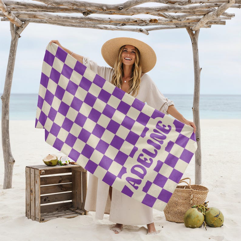 Op de afbeelding: Een strandlaken met een paars en wit dambordpatroon. De naam "ADELINE" is in paars op de handdoek gedrukt. De sc&egrave;ne speelt zich af op een strand.