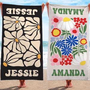 K&ouml;nnte beinhalten: Zwei Strandt&uuml;cher werden gezeigt. Eines ist schwarz mit gro&szlig;en G&auml;nsebl&uuml;mchenmustern und dem Namen "JESSIE" in Wei&szlig;. Das andere ist beige, mit bunten Blumen und dem Namen "AMANDA" in Gr&uuml;n. Beide werden an einem Strand gezeigt.