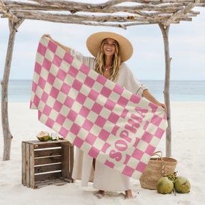 Op de afbeelding: Een roze en witte geblokte strandlaken met de naam "SOPHIA" in roze gedrukt. De handdoek wordt vastgehouden door een persoon op een zandstrand. Een houten kist, een rieten mand en kokosnoten zijn ook zichtbaar.