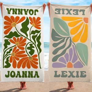 K&ouml;nnte beinhalten: Zwei Strandt&uuml;cher mit Blumendesigns und Namen. Ein Handtuch hat orangefarbene Blumen, gr&uuml;ne Bl&auml;tter und den Namen "JOANNA" in Gr&uuml;n. Das andere hat abstrakte Blumen in Gelb, Lila und Gr&uuml;n mit dem Namen "LEXIE".