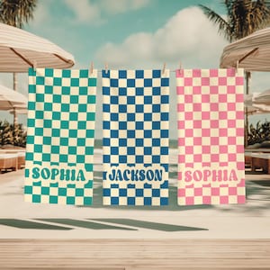 Op de afbeelding: Drie gepersonaliseerde strandlakens hangen aan een waslijn. De handdoeken hebben een dambordpatroon in teal, blauw en roze. De teal en roze handdoeken hebben de naam "SOPHIA" erop gedrukt, terwijl de blauwe handdoek "JACKSON" heeft.