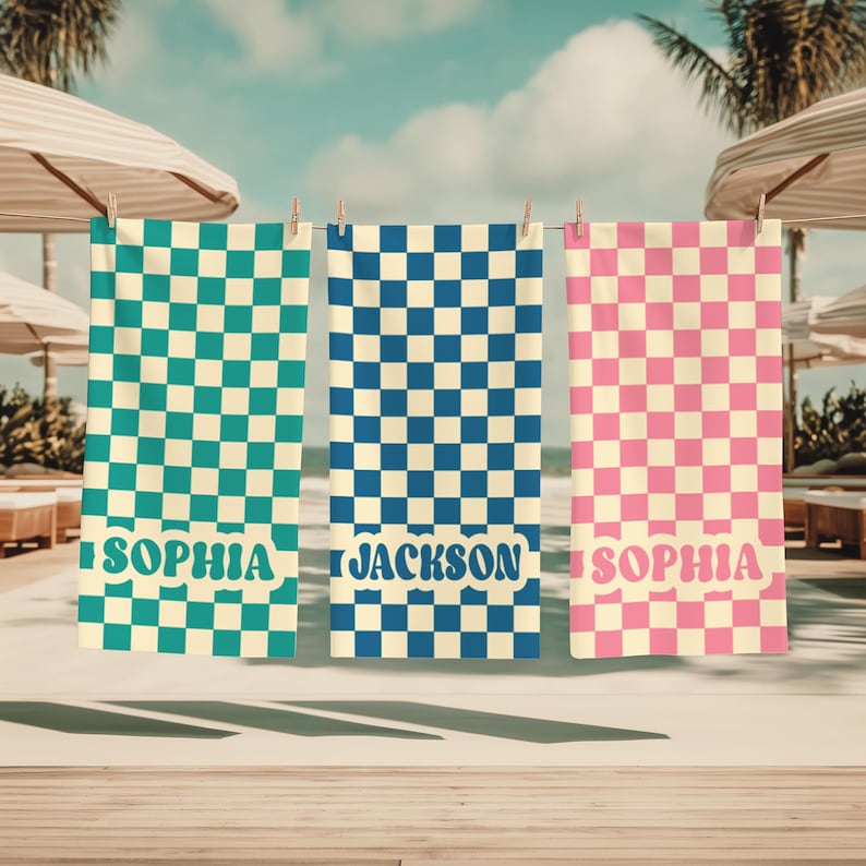 Op de afbeelding: Drie gepersonaliseerde strandlakens hangen aan een waslijn. De handdoeken hebben een dambordpatroon in teal, blauw en roze. De teal en roze handdoeken hebben de naam "SOPHIA" erop gedrukt, terwijl de blauwe handdoek "JACKSON" heeft.