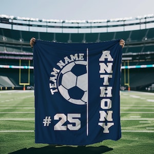 Könnte beinhalten: Eine dunkelblaue rechteckige Decke mit einer weißen Grafik eines Fußballs in der Mitte. Der Text "TEAM NAME" ist über dem Ball gebogen, "# 25" ist unten links aufgedruckt und der Name "ANTHONY" ist vertikal in weißer Schrift zu sehen.