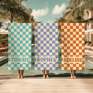 Op de afbeelding: Drie strandlakens met dambordpatronen in turquoise, lavendel en oranje. Elk laken heeft een naam onderaan afgedrukt: "TOMMY", "SOPHIA" en "ADELINE". De lakens worden voor een zwembad gehouden.