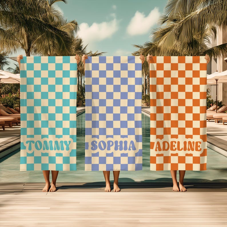Op de afbeelding: Drie strandlakens met dambordpatronen in turquoise, lavendel en oranje. Elk laken heeft een naam onderaan afgedrukt: "TOMMY", "SOPHIA" en "ADELINE". De lakens worden voor een zwembad gehouden.