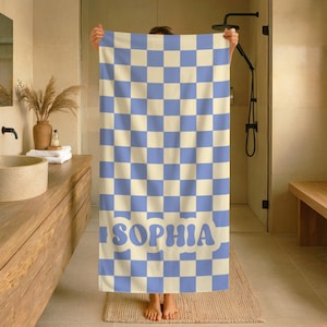 Op de afbeelding: Een blauw en cr&egrave;me geruite badhanddoek met de naam "SOPHIA" in blauwe blokletters. De handdoek wordt in een badkamer gehouden en toont een modern design. De afmetingen van de handdoek zijn niet zichtbaar.
