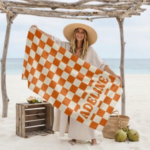 Op de afbeelding: Oranje en cr&egrave;mekleurige geblokte strandlaken met de naam "ADELINE" in oranje gedrukt. De handdoek wordt door een persoon op een strand omhoog gehouden. Een houten kist, een rieten mand en kokosnoten zijn ook zichtbaar.