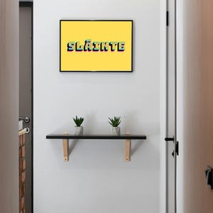 May include: A white wall with a yellow framed picture that says "Sl&aacute;inte" in colourful letters. There is a black shelf with two small green plants on it.