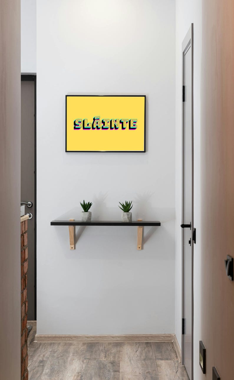 May include: A white wall with a yellow framed picture that says "Sl&aacute;inte" in colourful letters. There is a black shelf with two small green plants on it.