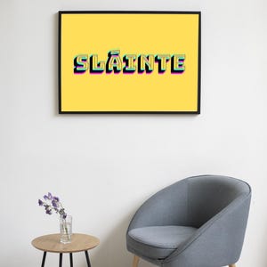 May include: A framed print with a yellow background and the word "Sl&aacute;inte" in a colourful, layered font. The print is hanging on a white wall above a grey armchair and a small wooden table with a vase of purple flowers.