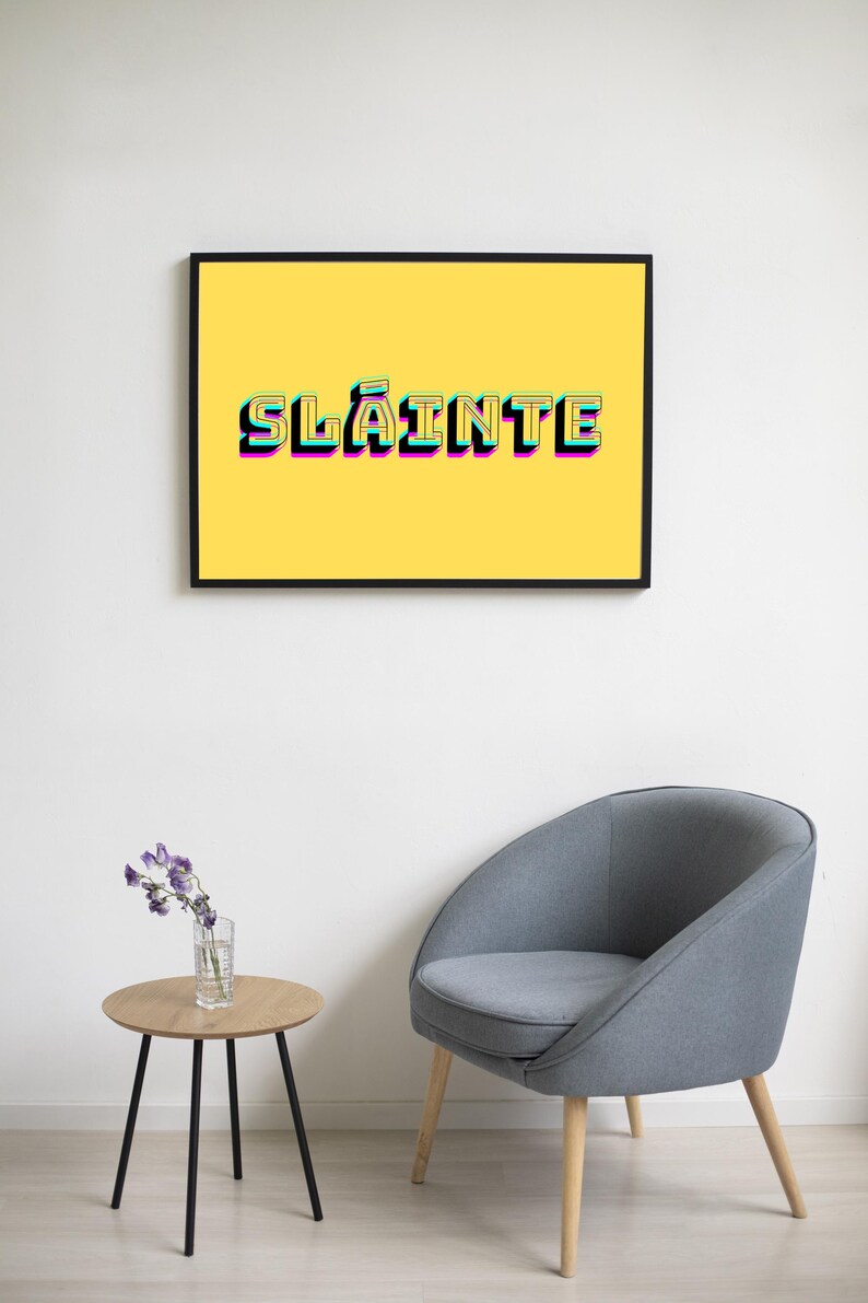 May include: A framed print with a yellow background and the word "Sl&aacute;inte" in a colourful, layered font. The print is hanging on a white wall above a grey armchair and a small wooden table with a vase of purple flowers.