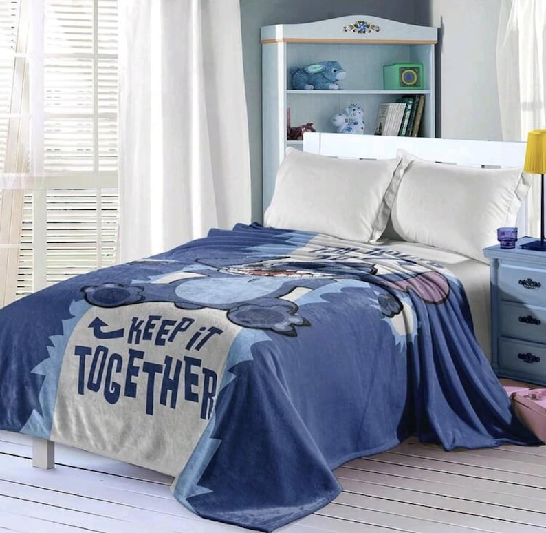 May include: A blue fleece blanket with a cartoon character design. The blanket features the text "KEEP IT TOGETHER" in white and blue. The blanket is draped over a bed with white pillows.