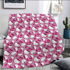 May include: Pink throw blanket featuring a repeating pattern of white Hello Kitty faces with pink bows. The blanket is draped over a light-colored sofa. A framed picture and a cactus in a blue pot are visible in the background.