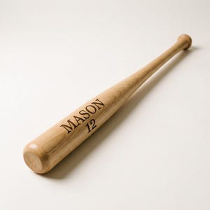 May include: A light brown wooden baseball bat with a smooth, polished finish. The name MASON and the number 12 are engraved in a dark, sans-serif font on the barrel of the bat. The bat features a rounded knob at the end of the handle.