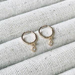 May include: A pair of gold-colored hoop earrings with a small, diamond-shaped charm hanging from each. The earrings are displayed on a textured, light gray surface.