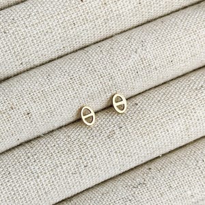 May include: A pair of small, gold-colored stud earrings. Each earring features a stylized, oval-shaped design with a horizontal bar across the center. The earrings are displayed on a textured, off-white fabric.