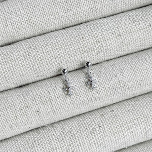 May include: A pair of silver-colored stud earrings. Each earring features a small, round stud with a dangling cross design. The earrings are displayed on a textured, light-colored fabric background.