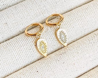 May include: A pair of gold-colored hoop earrings. Each earring features a teardrop-shaped charm with a white enamel inlay and a geometric design. The earrings are displayed on a textured, off-white surface.