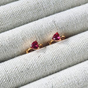 May include: A pair of gold heart-shaped earrings with red cubic zirconia stones.
