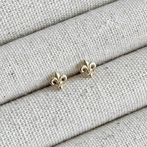 May include: A pair of small, gold-coloured fleur-de-lis stud earrings. The earrings are presented on a textured, light-coloured fabric. The fleur-de-lis design is a stylized lily, a symbol of French royalty.